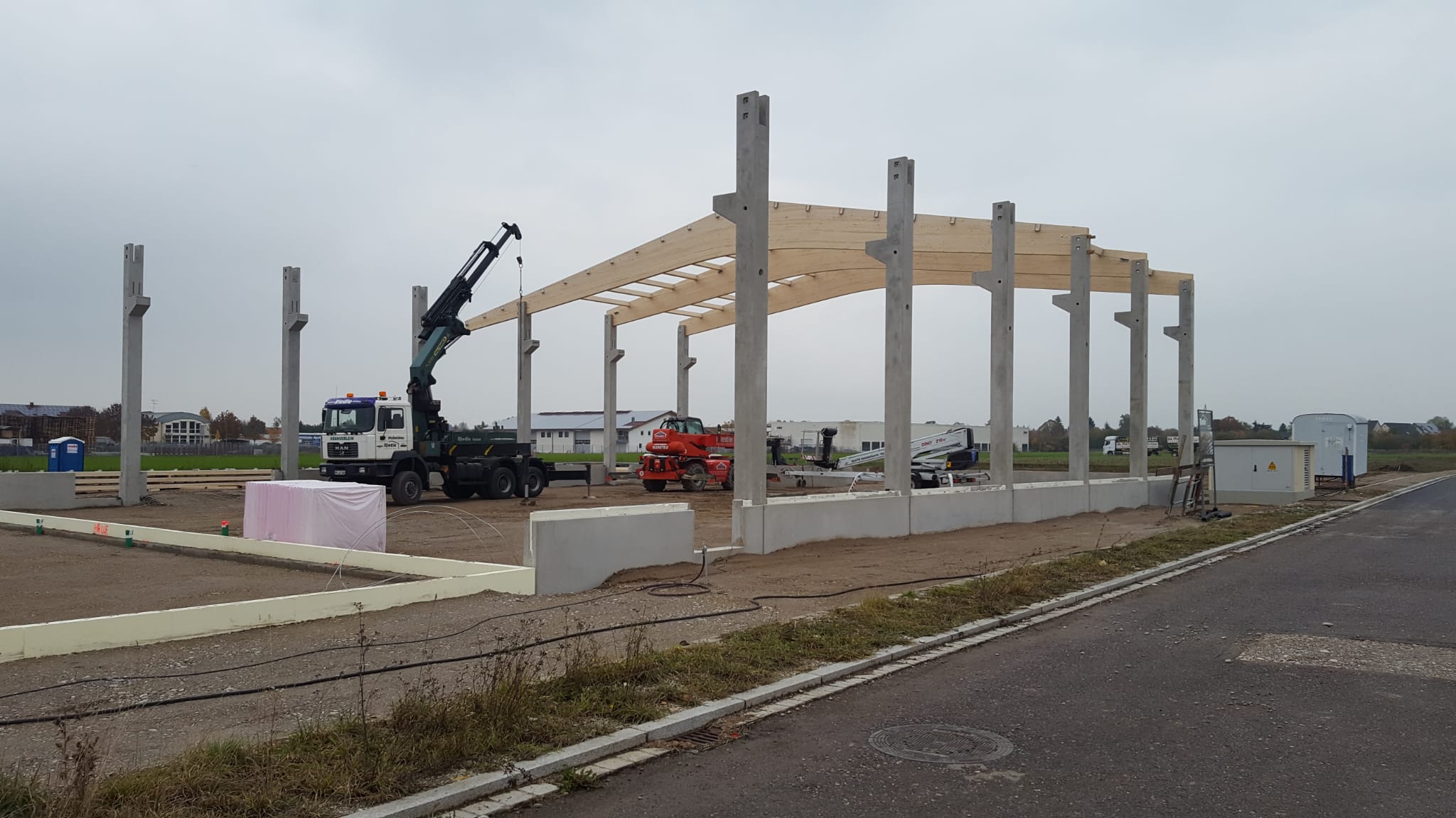 Rohbau: der erste Binder aus Brettschichtholz liegt auf den Betonstützen, ein Autokran daneben