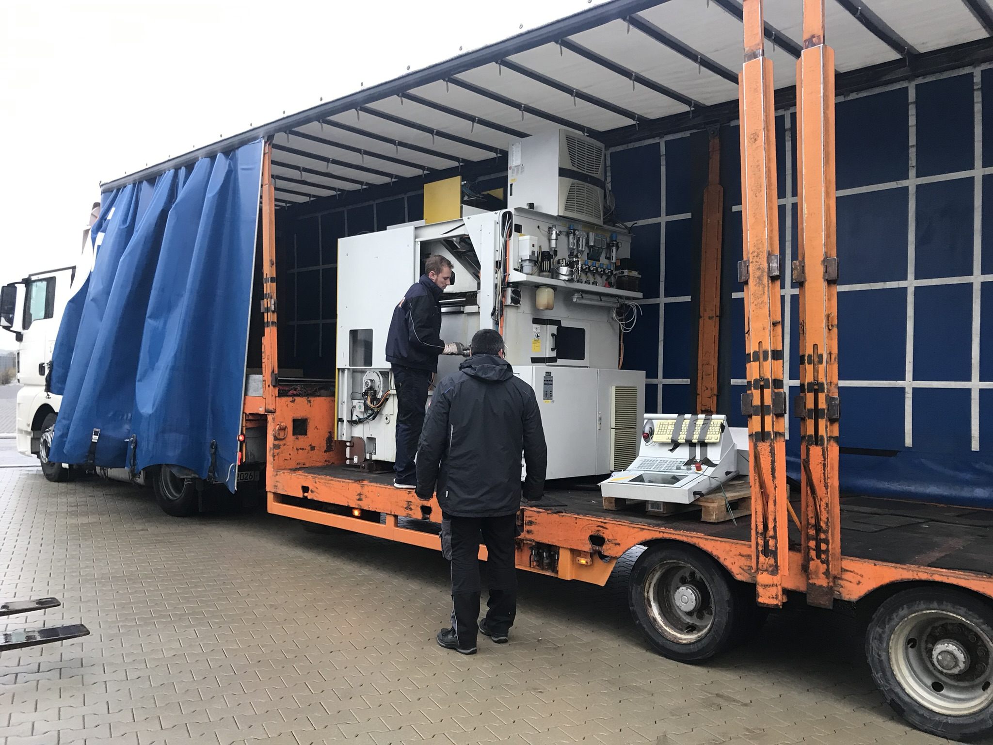 Zwei Mitarbeiter lösen die Maschine auf dem Tieflader, das Bedienpult steht daneben auf einer Palette