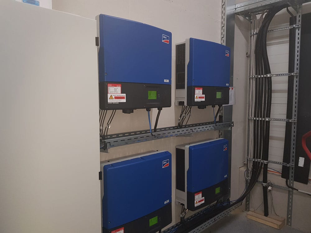 Vier blaue Wechselrichter nebeneinander an der Wand des Technikraums, rechts die Kabeltrasse