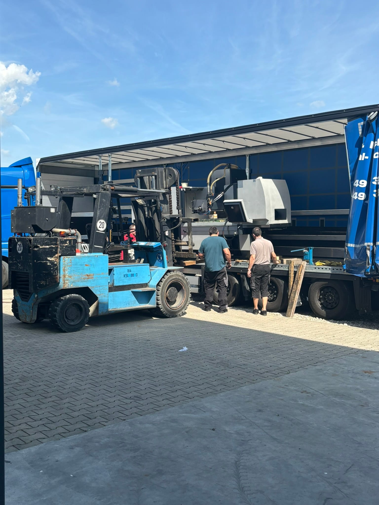 Der blaue Schwerlaststapler ist vor den Auflieger gefahren, zwei Mitarbeiter richten die Gabeln aus
