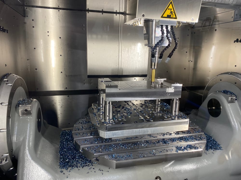Formplatte im Bearbeitungszentrum, auf Bolzen über der Aufspannplatte abgestützt, Fräser im Eingriff, blaue Späne auf dem Schwenkrundtisch