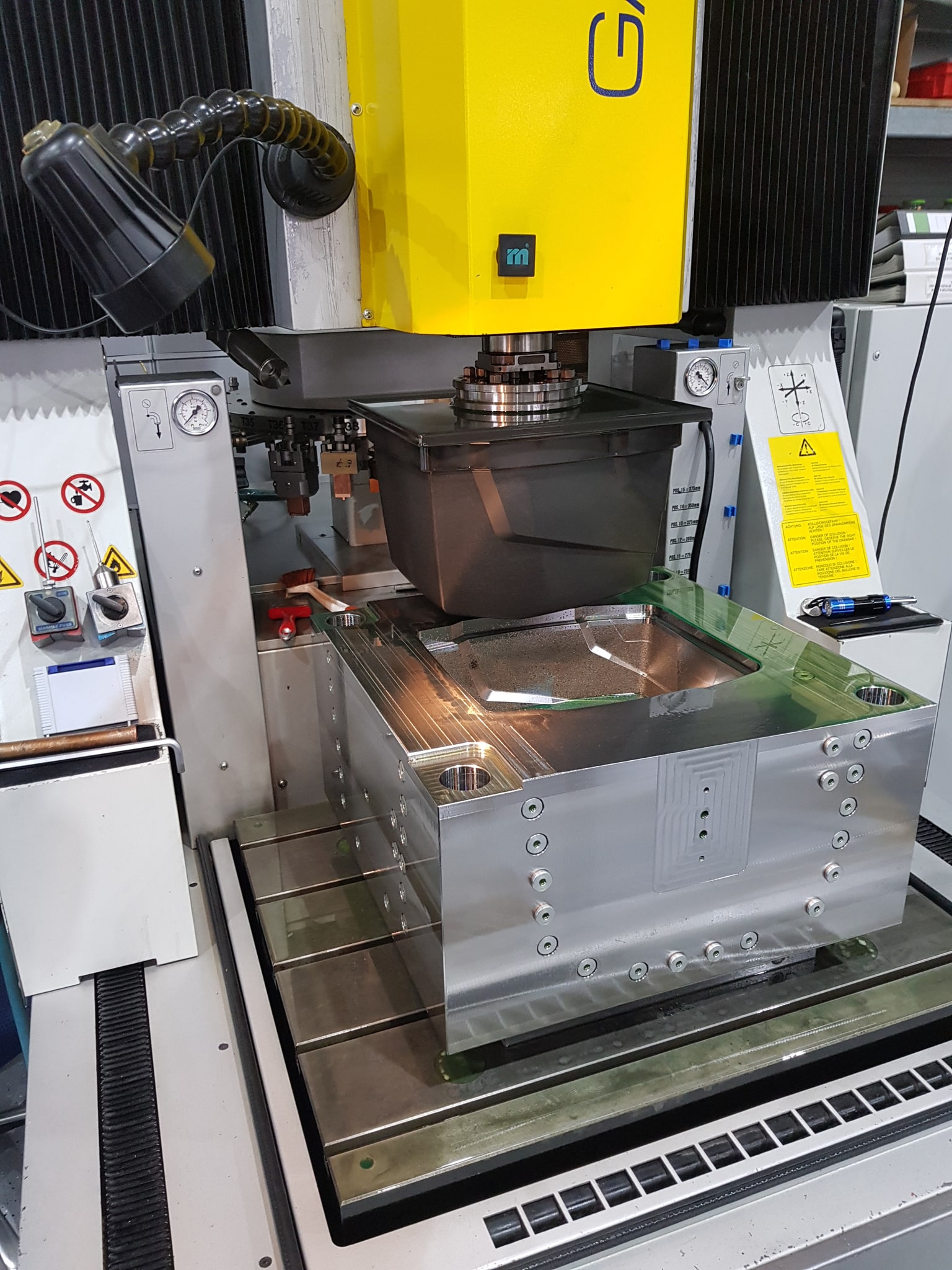 Formplatte auf dem Tisch der Senkerodiermaschine, darüber die große Graphitelektrode am Maschinenkopf, in der Platte die blanke Kavität des Sichtteils