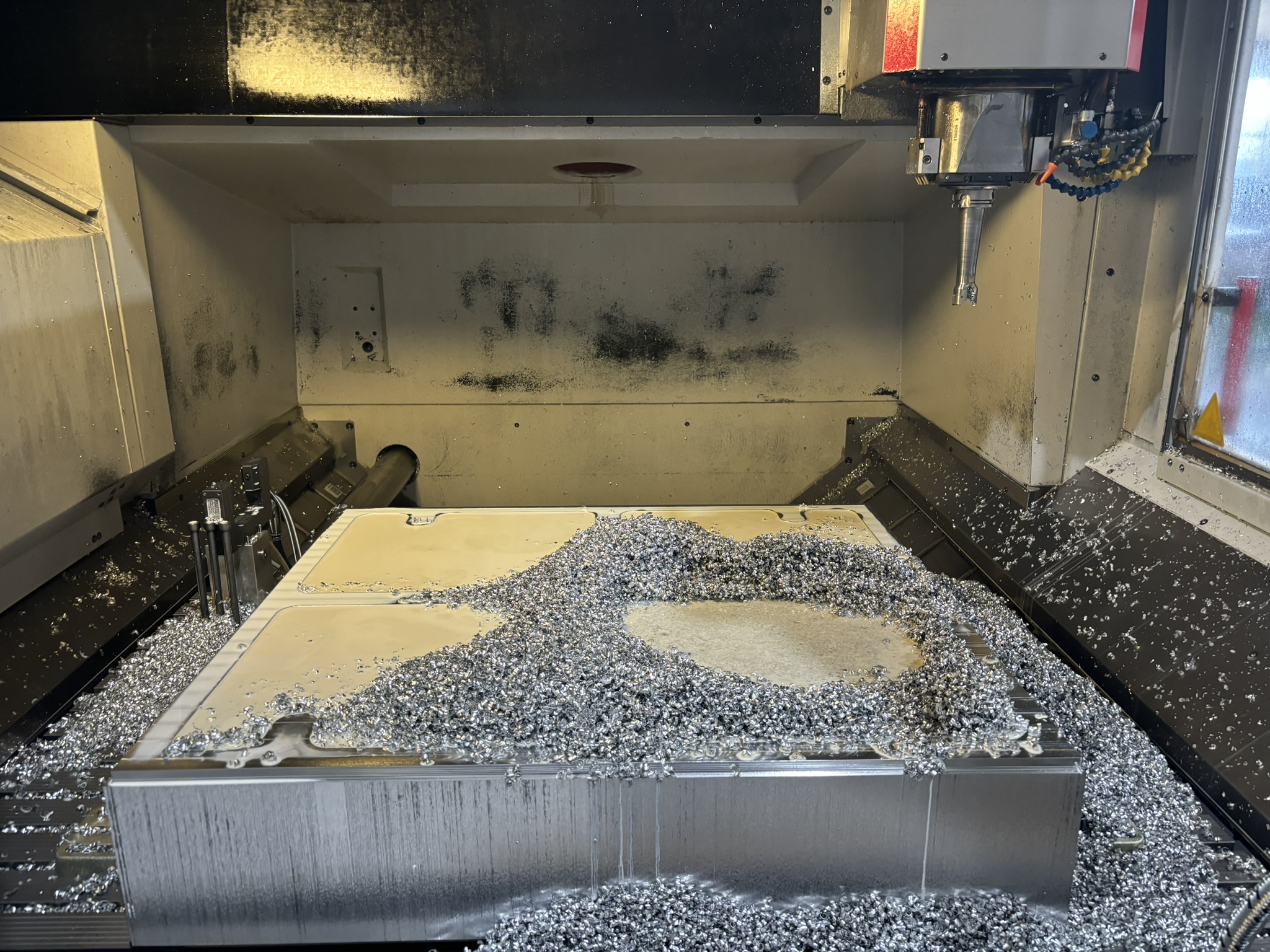 Großes Aluminiumbauteil im Bearbeitungszentrum, in der Fläche ausgefräste Taschen, darauf und ringsum Berge von Frässpänen, darüber der Fräser in der Spindel